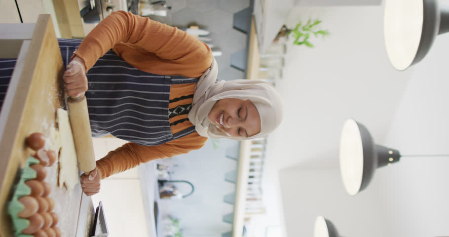 Vertical video of happy biracial woman in hijab baking in kitchen, rolling dough with rolling pin. Happiness, health, inclusivity and domestic life.