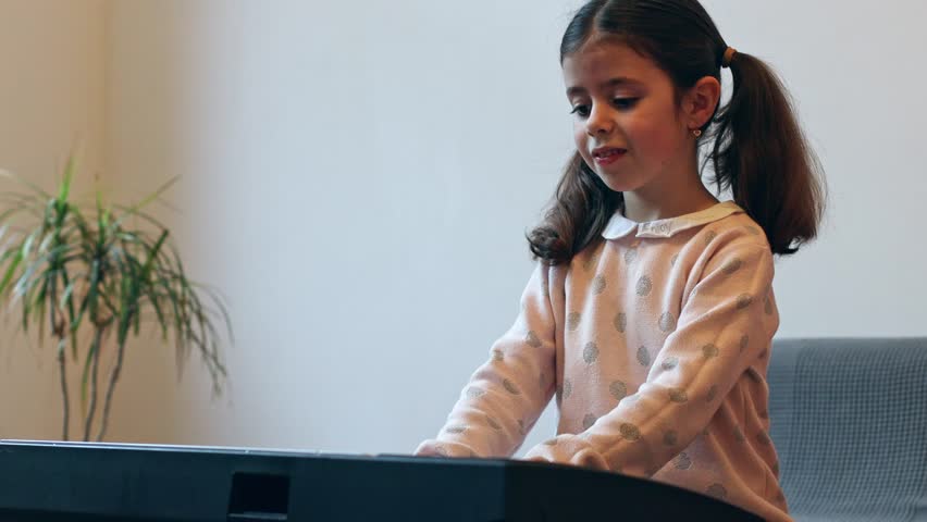 Beautiful little caucasian brunette girl with two ponytails plays the electric piano and sings while sitting on the sofa in the room, top view close-up.Music education concept.
