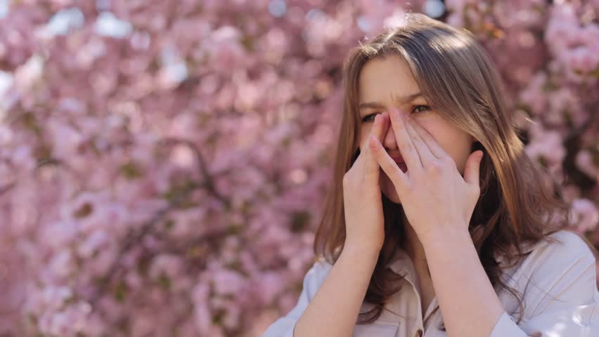 Irritated caucasian young woman standing near blooming sakura tree and scratching itchy eyes during spring time outdoors. Allergic reaction to pollen. For Advertisement of a medicinal product | Shutterstock HD Video #1099738953