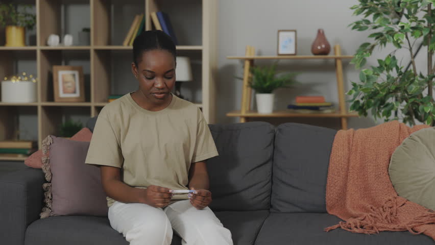 Sad african american woman looking at negative pregnancy test with disappointed expression while sitting on cozy couch at home. Concept of infertility problem. | Shutterstock HD Video #1099741159