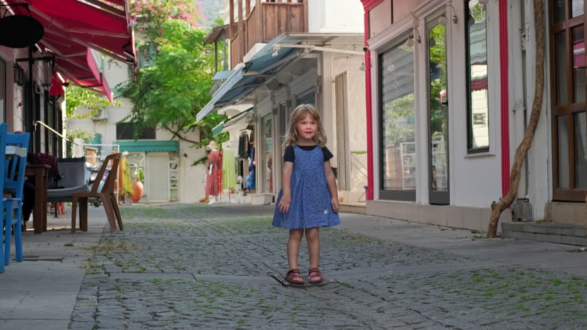 curly-haired blonde traveler girl in blue dress stands in middle of small pedestrian street in old town. Portrait of cheerful child with white flower in her hand during day on trip. look around street