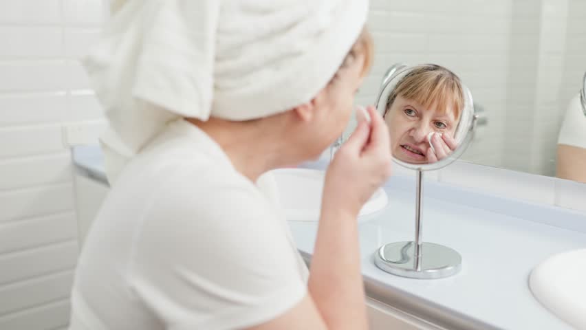 Gorgeous mid age older adult 50 years old blonde woman wears bathrobe in bathroom applying nourishing antiage face skin care cream treatment, looking at mirror doing daily morning beauty routine. Royalty-Free Stock Footage #1099744865