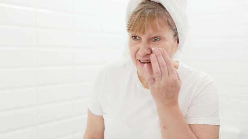 Gorgeous mid age older adult 50 years old blonde woman wears bathrobe in bathroom applying nourishing antiage face skin care cream treatment, looking at mirror doing daily morning beauty routine. Royalty-Free Stock Footage #1099744873