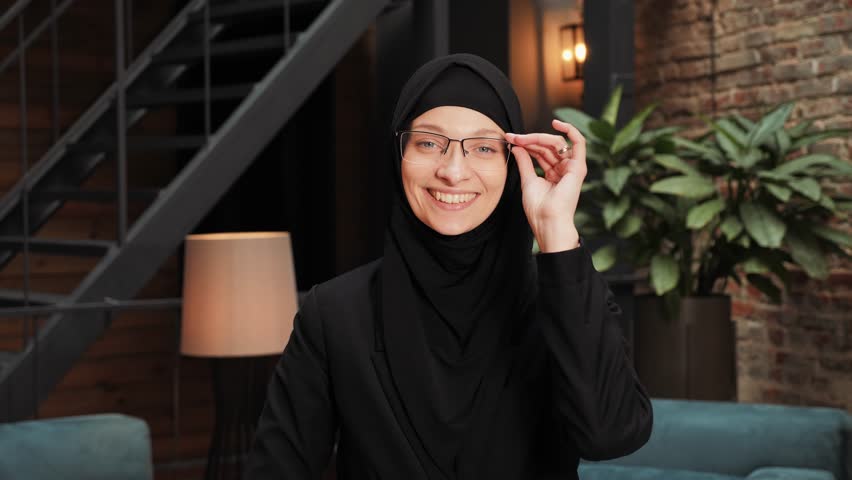 Portrait of beautiful pretty Arab muslim woman in black traditional hijab looking straight to camera and smiling in modern loft office. Close up of female pretty face with smile. Arabian headscarf.