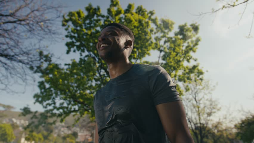 Young black man in t-shirt smiling while taking a break and drinking water by trees in park Royalty-Free Stock Footage #1099747665