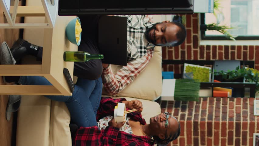 Vertical video: Life partners watching television together at home, eating noodles with chopsticks and browsing internet website on laptop. Cheerful modern couple having fun with tv show, drinking | Shutterstock HD Video #1099747777