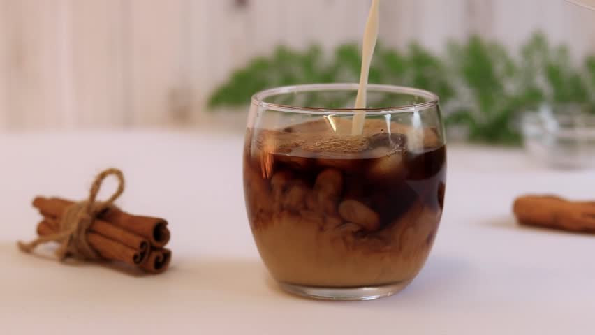Iced coffee with milk, a refreshing invigorating drink. Milk is poured into a cup with coffee on the background of a bright kitchen. Coffee with milk | Shutterstock HD Video #1099750897