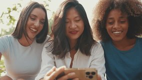 Close-up of happy multiethnic young women watching video on cellphone on summer day outdoors. Group of friends use smartphone, watch videos, social networks