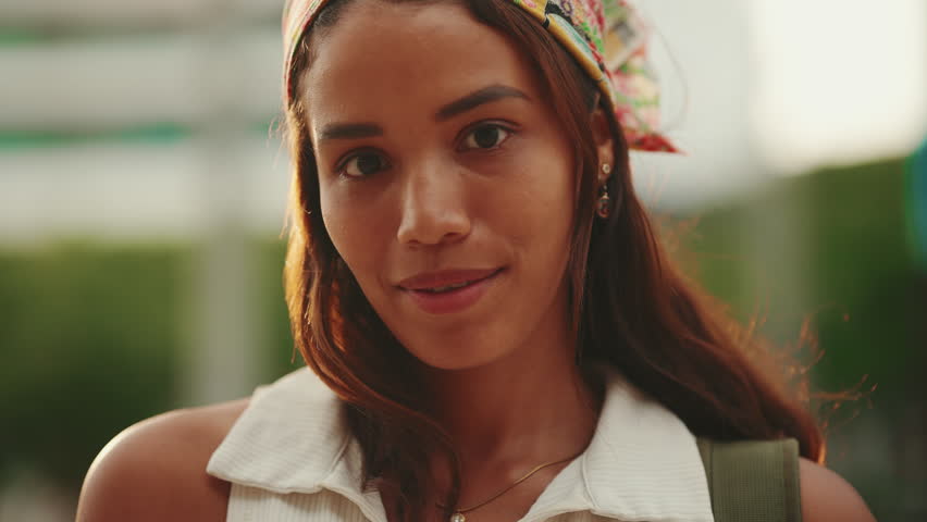 Clouse-up, cute tanned woman with long brown hair wearing white top and yellow bandana posing on camera. Smiling girl looking at the camera. Backlight