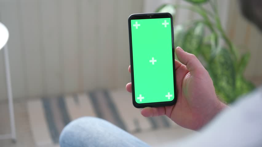Young man sitting at home holding smartphone green mock-up screen in hand. Male person using chroma key mobile phone. Vertical mode. Touching, swiping display, tapping, surfing Internet social media | Shutterstock HD Video #1099759951