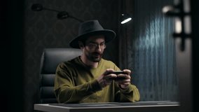 Man in Hat play video game on smartphone sitting at home.Portrait of young guy holding mobile phone enjoying gaming indoors.Celebrating victory game concept. High quality 4k footage