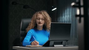 Curly woman learning online using laptop and notepad sitting at home.Young girl make notes with serious face in dark room.Lady doing education english online . High quality 4k footage