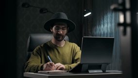 Young Bearded Man learning online using laptop sitting at desk in dark room.Smart guy wear hat make video call doing online education for students home remote.Male work remotely class lesson concept
