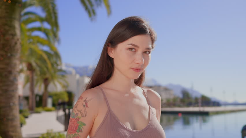 Close-up portrait of joyful young asian woman with dark hair and tattoo standing alone in seaside city smiling looking at camera. People and style concept. Palm trees ocean and mountains in background | Shutterstock HD Video #1099765171