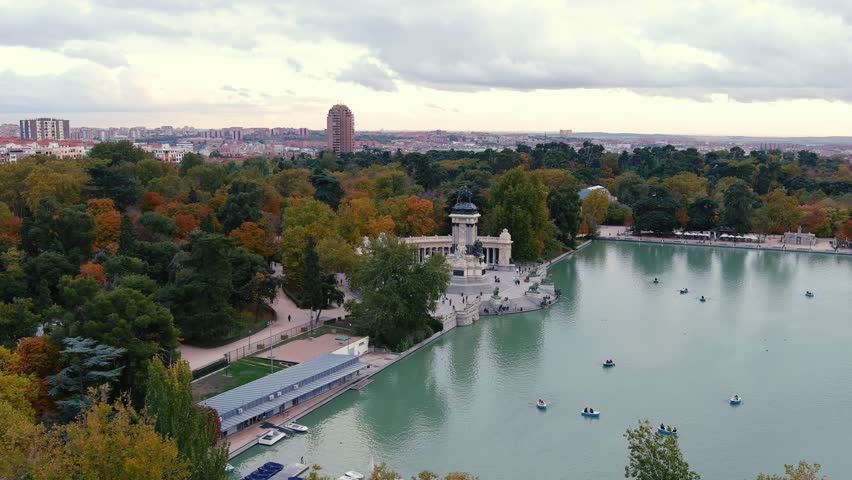 The drone is rotating around Monumento Alfonso XII in Parque del Buen Retiro with autumn color trees and people rowing in the pond in Madrid Spain Aerial Drone Footage 4K