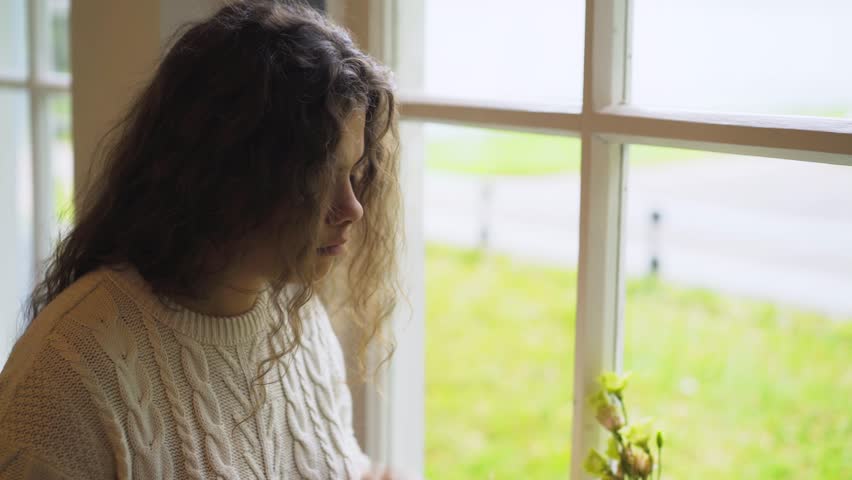 Portrait young concentrated female florist in white sweater chooses and arranges flowers to make bouquet in daylight opposite window, taking part in interesting floral masterclass