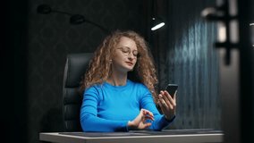 Young Curly Woman make video call using phone sitting on a chair at the table.Lady doing online communication looking at smartphone.Young smiling girl doing meeting with friends in dark room. High