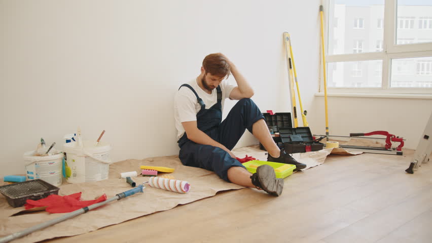 Confused annoyed tired man sitting on floor with rolls of wallpaper, instruments for renovation apartment room. Repair home concept. Pensive tired man looking away while making repair. Royalty-Free Stock Footage #1099784349