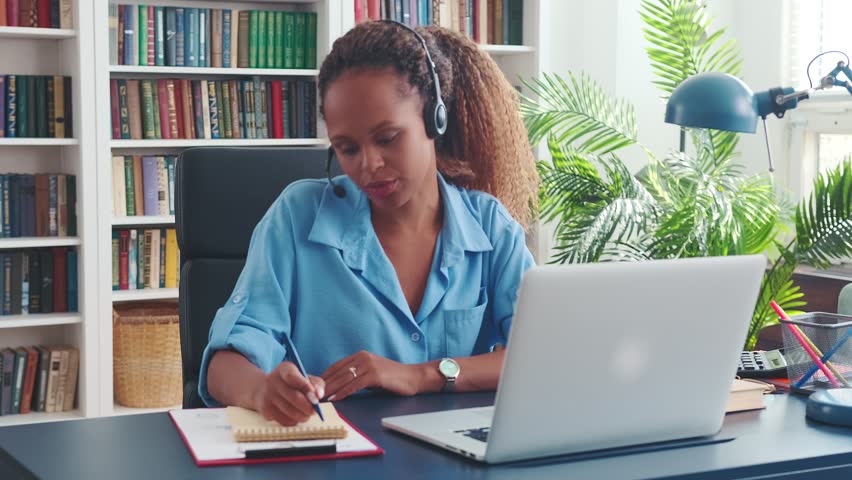 Young attractive casual African American woman in headphones with microphone participates in morning online meeting of remote employees of company discussing work plan sits at table in office | Shutterstock HD Video #1099788807