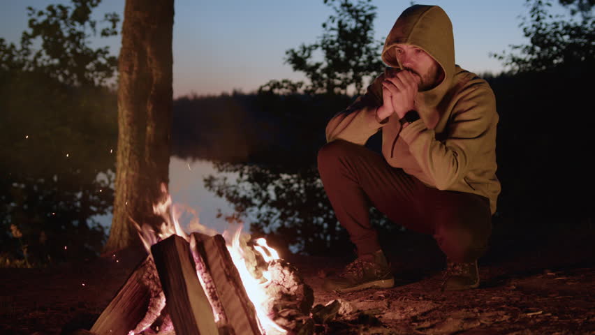 Young bearded hiker squats at campfire warming palms over flame and breathing in hands