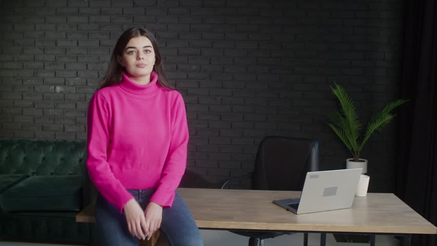 A business lady in a pink sweater smiles and crosses her arms. Brunette girl sitting on the table. Remote work from the studio. | Shutterstock HD Video #1099793469