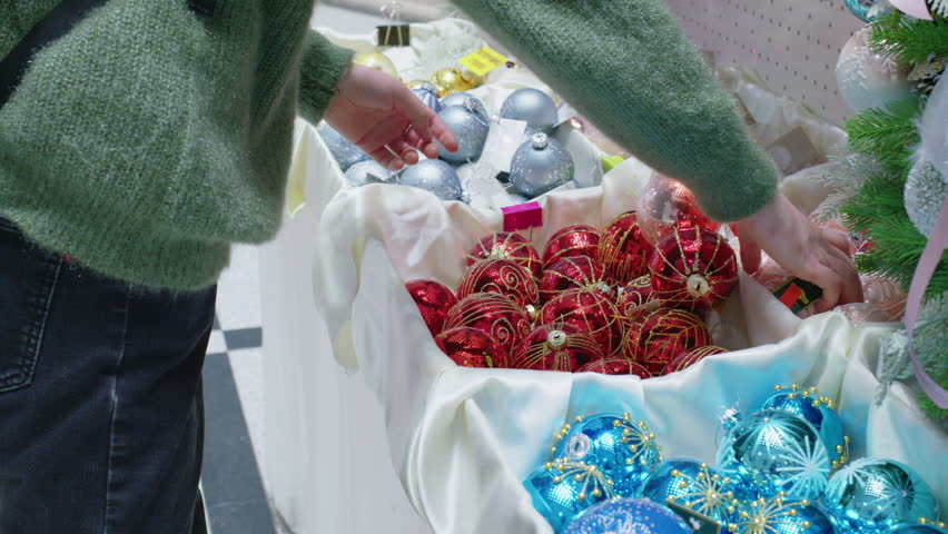 Faceless girl chooses balls, baubles for xmas tree in store. Unrecognizable woman is looking at assortment of Christmas ornament, decorations in shop to buy something for gift. Holiday shopping