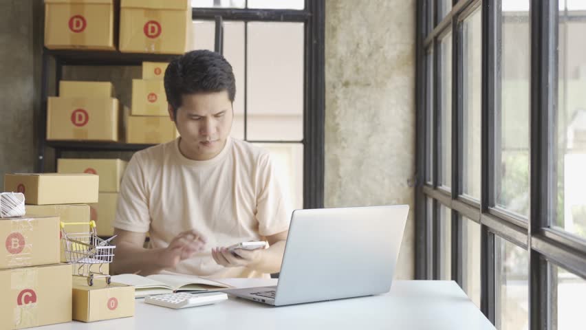 Young Asian businessman in T-shirt among parcels Working in a home office with a laptop taking orders, name looks like doing startup business for parcel box delivery, SME online marketing. | Shutterstock HD Video #1099795417
