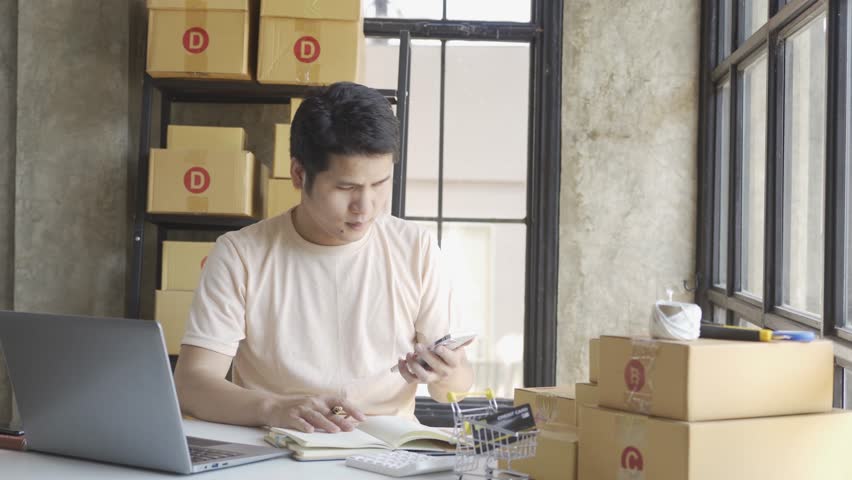 Young Asian businessman in T-shirt among parcels Working in a home office with a laptop taking orders, name looks like doing startup business for parcel box delivery, SME online marketing. | Shutterstock HD Video #1099795419