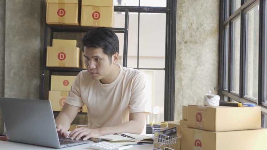 Young Asian businessman in T-shirt among parcels Working in a home office with a laptop taking orders, name looks like doing startup business for parcel box delivery, SME online marketing. | Shutterstock HD Video #1099795421