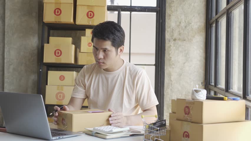 Young Asian businessman in T-shirt among parcels Working in a home office with a laptop taking orders, name looks like doing startup business for parcel box delivery, SME online marketing. | Shutterstock HD Video #1099795425