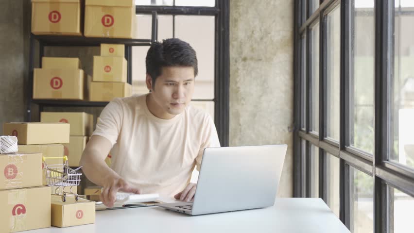 Young Asian businessman in T-shirt among parcels Working in a home office with a laptop taking orders, name looks like doing startup business for parcel box delivery, SME online marketing. | Shutterstock HD Video #1099795427