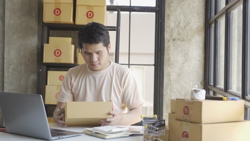 Young Asian businessman in T-shirt among parcels Working in a home office with a laptop taking orders, name looks like doing startup business for parcel box delivery, SME online marketing. | Shutterstock HD Video #1099795429