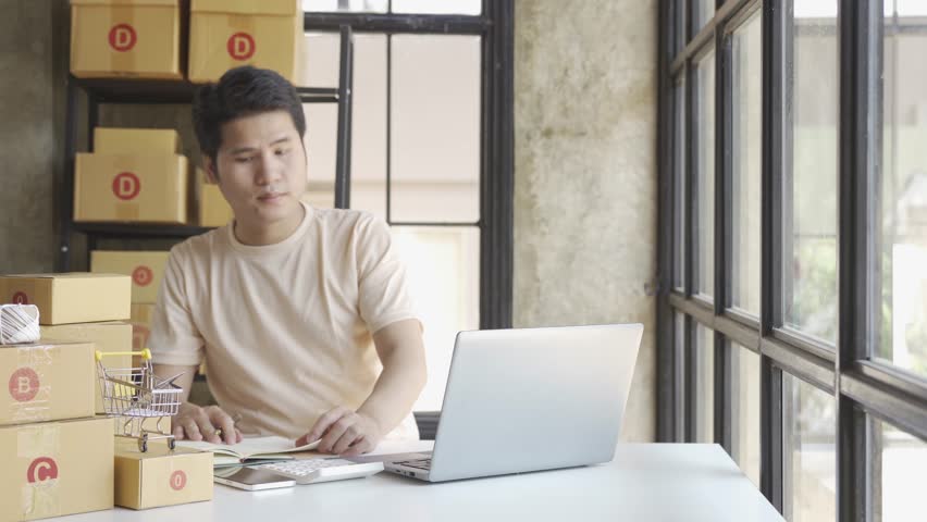 Young Asian businessman in T-shirt among parcels Working in a home office with a laptop taking orders, name looks like doing startup business for parcel box delivery, SME online marketing. | Shutterstock HD Video #1099795431
