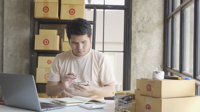 Young Asian businessman in T-shirt among parcels Working in a home office with a laptop taking orders, name looks like doing startup business for parcel box delivery, SME online marketing. | Shutterstock HD Video #1099795433