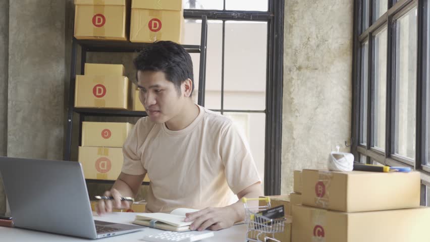 Young Asian businessman in T-shirt among parcels Working in a home office with a laptop taking orders, name looks like doing startup business for parcel box delivery, SME online marketing. | Shutterstock HD Video #1099795435