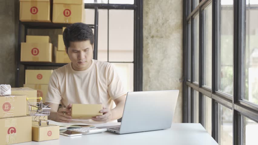 Young Asian businessman in T-shirt among parcels Working in a home office with a laptop taking orders, name looks like doing startup business for parcel box delivery, SME online marketing. | Shutterstock HD Video #1099795441