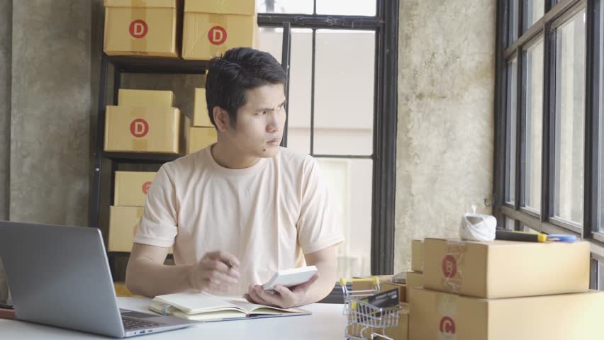 Young Asian businessman in T-shirt among parcels Working in a home office with a laptop taking orders, name looks like doing startup business for parcel box delivery, SME online marketing. | Shutterstock HD Video #1099795443