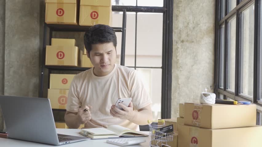 Young Asian businessman in T-shirt among parcels Working in a home office with a laptop taking orders, name looks like doing startup business for parcel box delivery, SME online marketing. | Shutterstock HD Video #1099795445