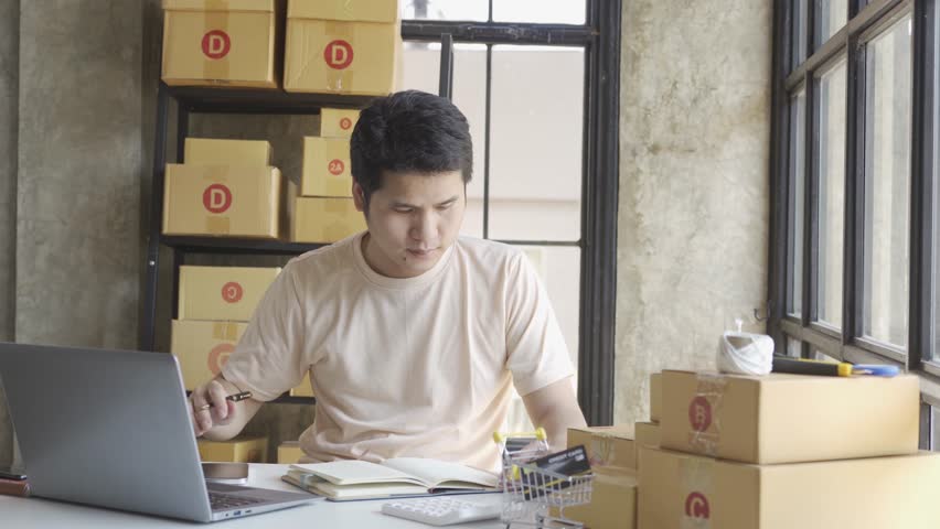 Young Asian businessman in T-shirt among parcels Working in a home office with a laptop taking orders, name looks like doing startup business for parcel box delivery, SME online marketing. | Shutterstock HD Video #1099795447