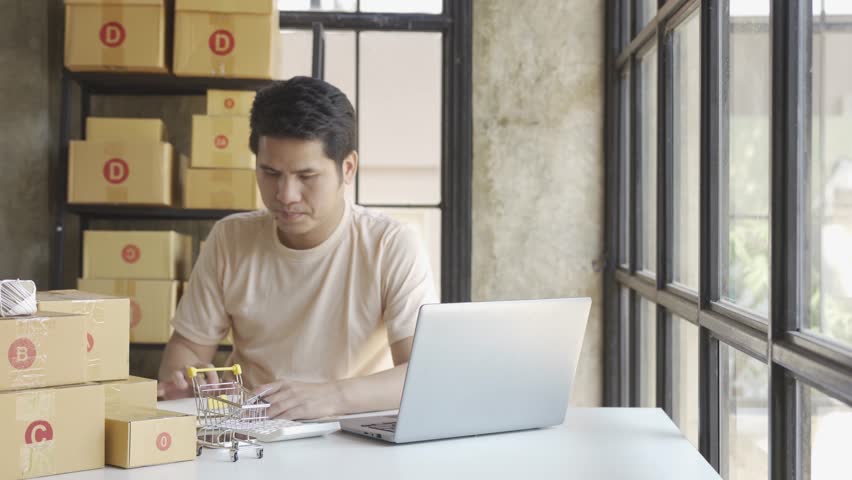 Young Asian businessman in T-shirt among parcels Working in a home office with a laptop taking orders, name looks like doing startup business for parcel box delivery, SME online marketing. | Shutterstock HD Video #1099795449
