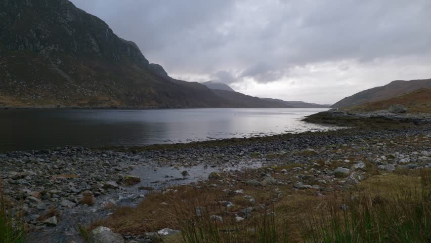 Water gently flows into a sea loch (Gleann Dubh) at low tide in the highlands of Scotland as moody dark clouds move across the sky behind a mountain.