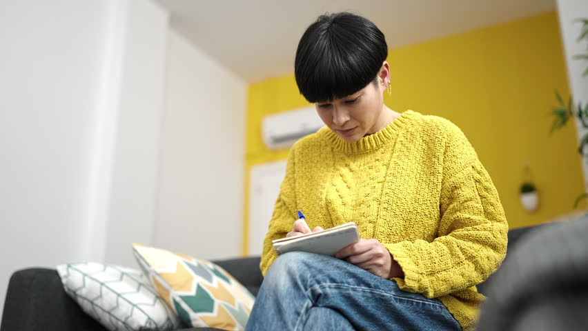 Young chinese woman writing on notebook sitting on sofa at home