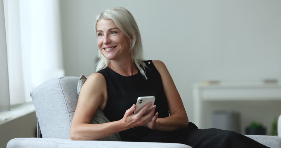 Attractive middle-aged woman relax on armchair using smartphone, resting at home chatting on-line, websurfing webpage, enjoy leisure on internet. Mature generation and modern wireless technology usage
