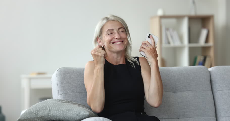 Mature 45s woman sitting on sofa staring at cellphone screen, read sms with pleasant news feels happy, get sell-out notice, lottery victory notification. Moment of win, advance, achievement, new apps Royalty-Free Stock Footage #1099811133