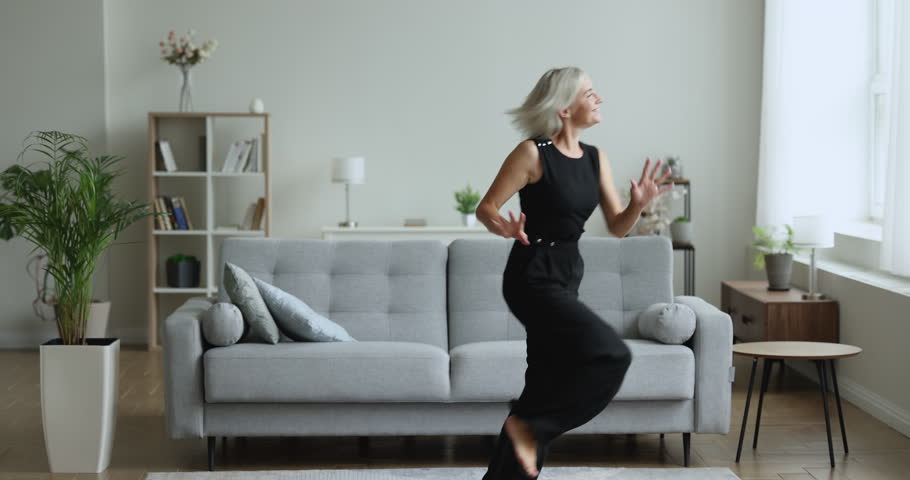Carefree mature woman having fun, running, jumping, moving in cozy warm living room feels happy, enjoy untroubled active weekend leisure alone at new own modern house. Tenancy, holiday, accommodation Royalty-Free Stock Footage #1099811153