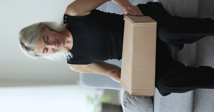 Client of reliable transport company, happy 50s woman sit on couch at home puts cardboard box on laps, smile enjoy process of received parcel unpack with ordered books for favourite hobby or education | Shutterstock HD Video #1099811187
