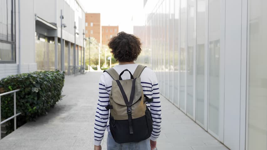 Student guy walking along university campus building. Happy and smiling, looking at the camera and going forward  Royalty-Free Stock Footage #1099816735