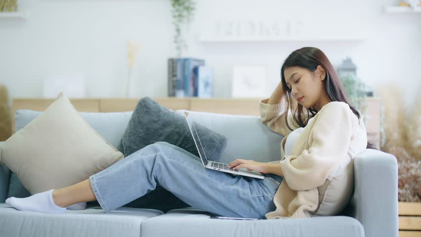 Young asian woman using computer laptop and mobile smartphone while seated on couch at home | Shutterstock HD Video #1099818105