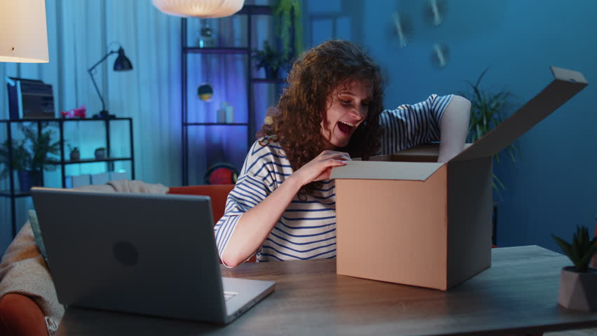 Happy surprised winner woman unpacking delivery parcel at home. Smiling satisfied girl shopper online shop customer opening cardboard box receiving purchase gift headphones by fast postal shipping | Shutterstock HD Video #1099818157