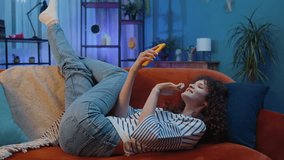 Young woman with curly hairstyle lying on sofa uses mobile phone smiles at night evening home. Girl texting share messages content on smartphone social media applications online watching relax movie